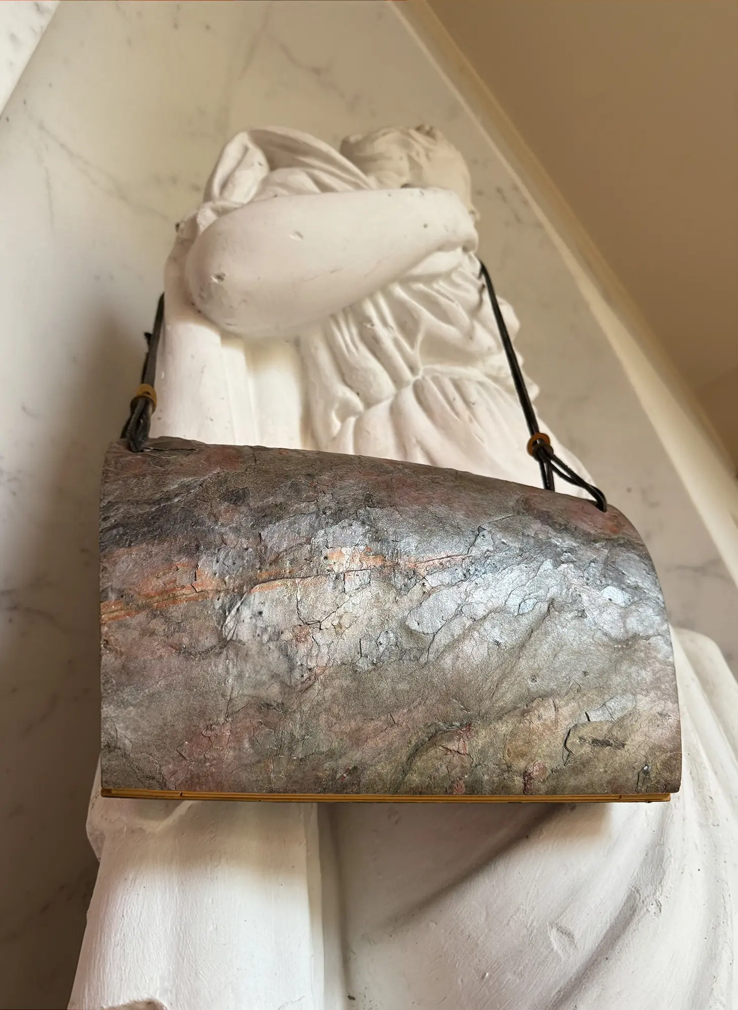 Pyroklast Abendtasche mit Schieferoberfläche und Lederträgern von einer weißen Statue getragen